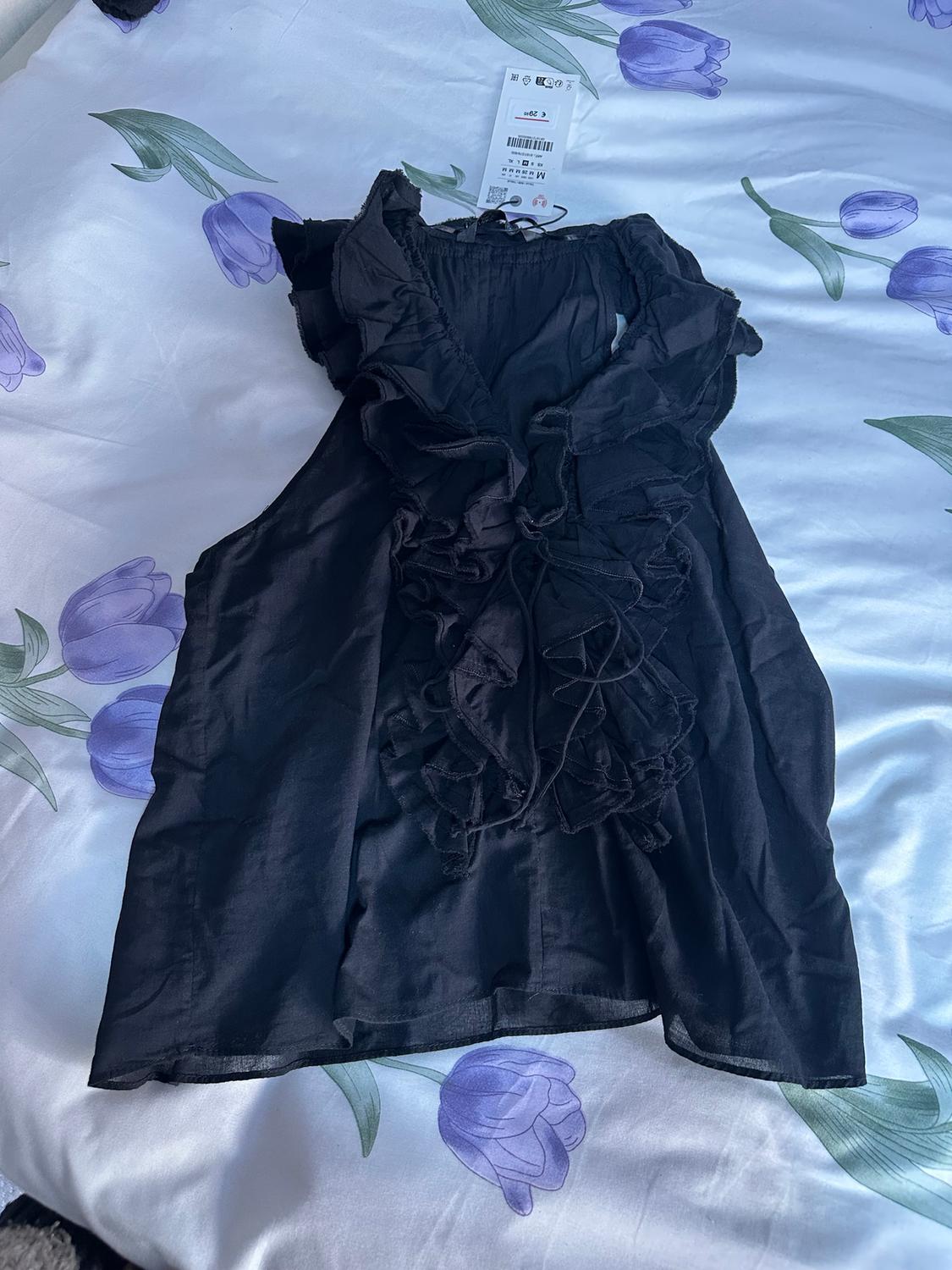 Black blouse with ruffles on a bed with purple floral sheets. Label visible at the top.