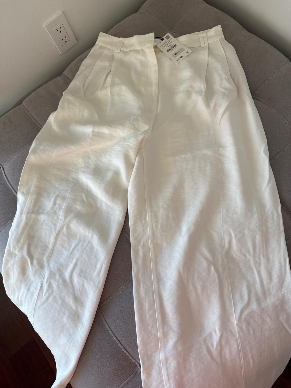 White linen trousers with pleats, visible tag, on a light grey sofa. Plug socket on the wall in the background.