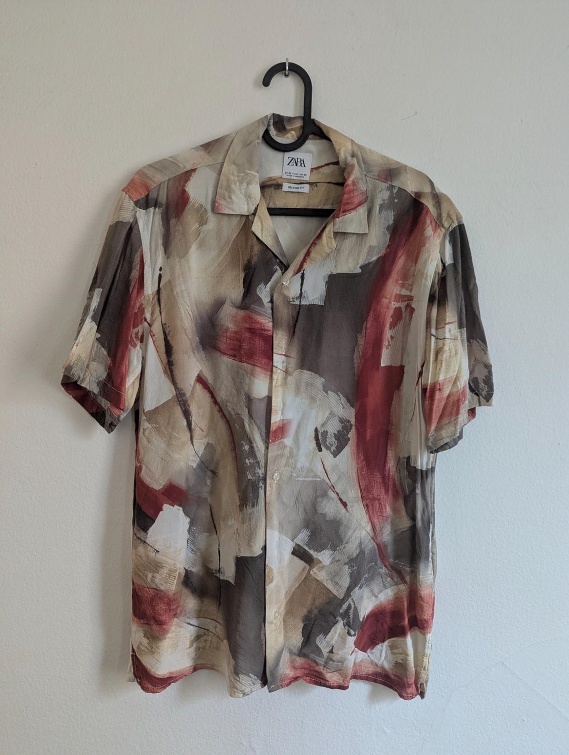 Short-sleeved shirt with an abstract print in beige, brown, and red tones, hanging on a black hanger. Visible label: Zara.