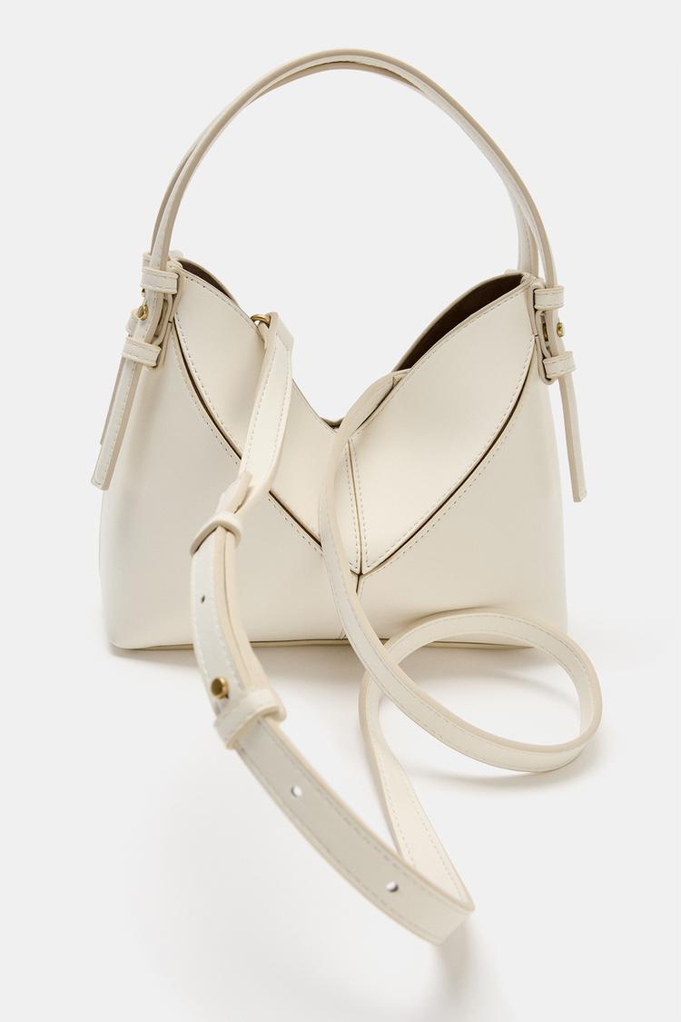 Bolso crema con asa superior y correa ajustable. Diseño estructurado con costuras visibles y detalles metálicos dorados.