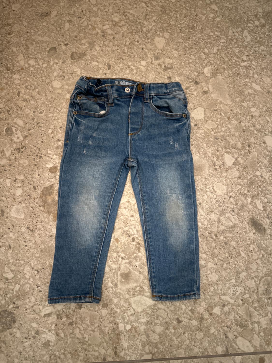 Blue denim jeans for children on a grey stone floor, with distressed details and a brown button.