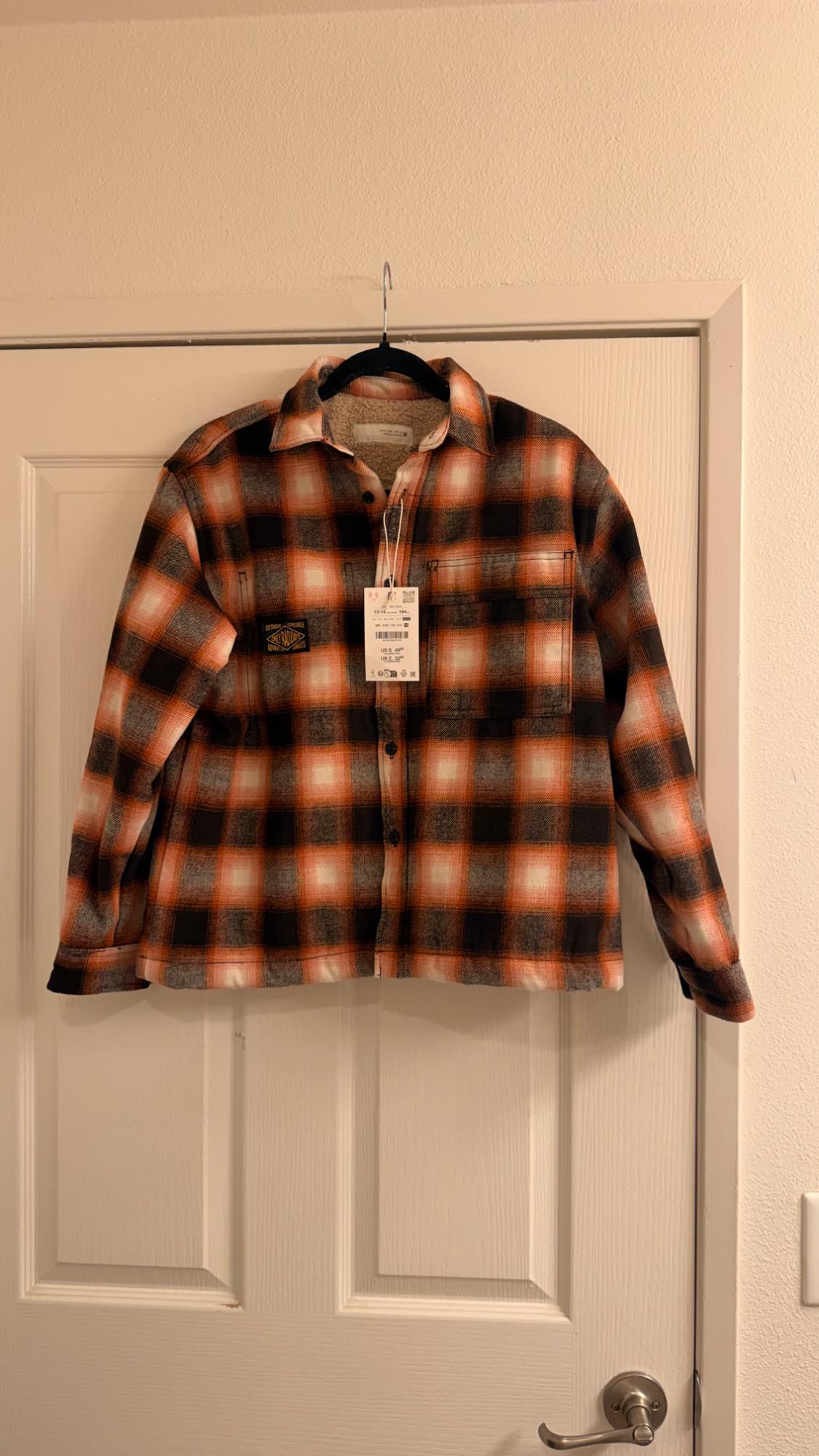 Orange, black, and white plaid shirt hanging on a white door. Casual style with a soft lining and an OUTDOOR EXPLORER patch.