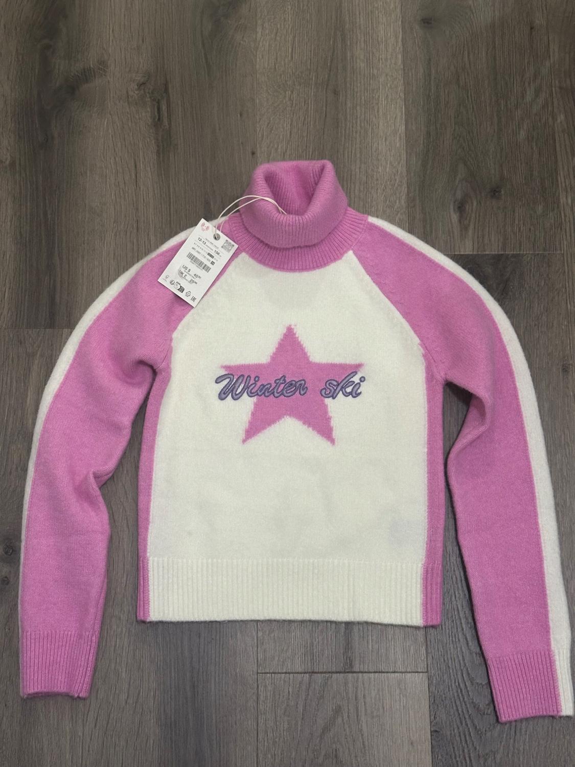 Pink and white sweater with a high collar, pink star, and "Winter ski" text on the front. Visible hang tag. Wooden background.