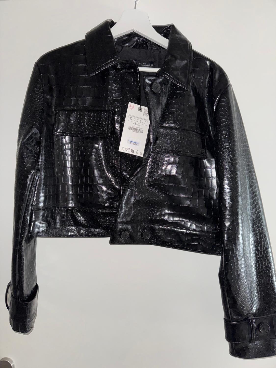 Black crocodile-effect jacket hanging on a white hanger, with a visible tag on the front.