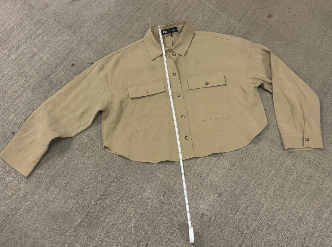 Long-sleeved beige shirt with front pockets, laid on the ground. A tape measure is aligned vertically over the shirt.