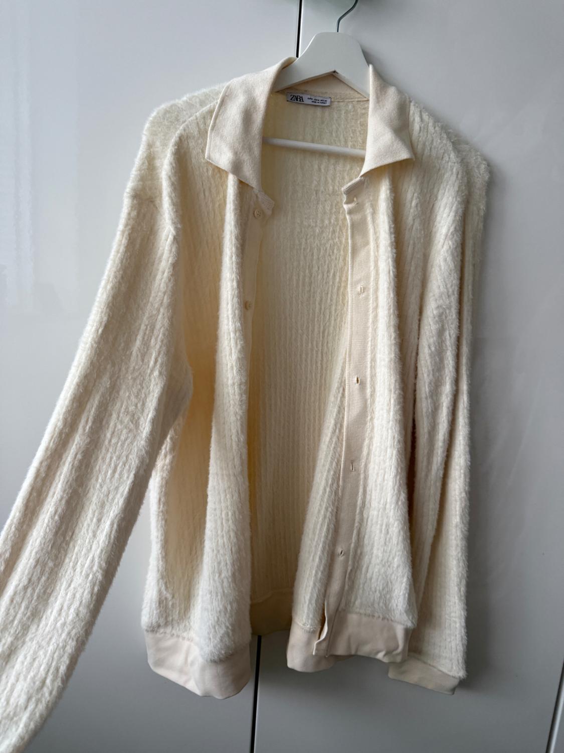 White knitted cardigan hanging on a white hanger, with a white wardrobe background. Visible label: "Zara".