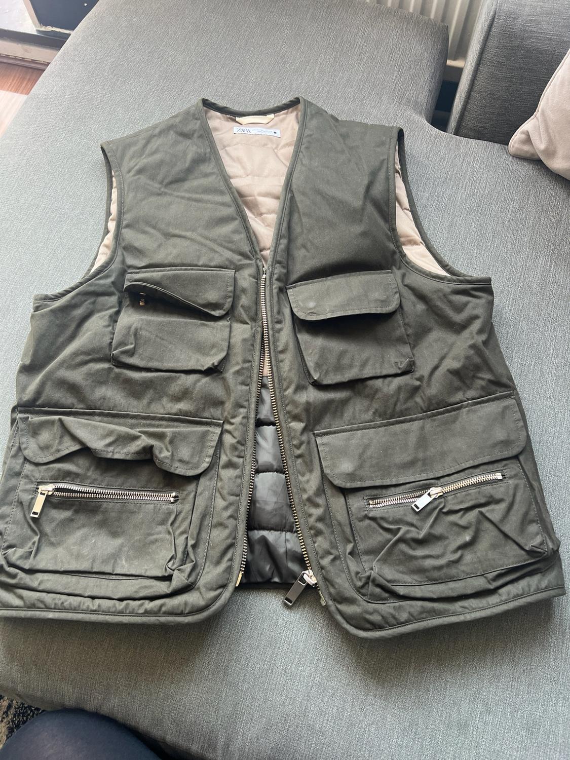 Olive green multi-pocket gilet with a zip and beige quilted lining, on a grey sofa. Text: Zara.