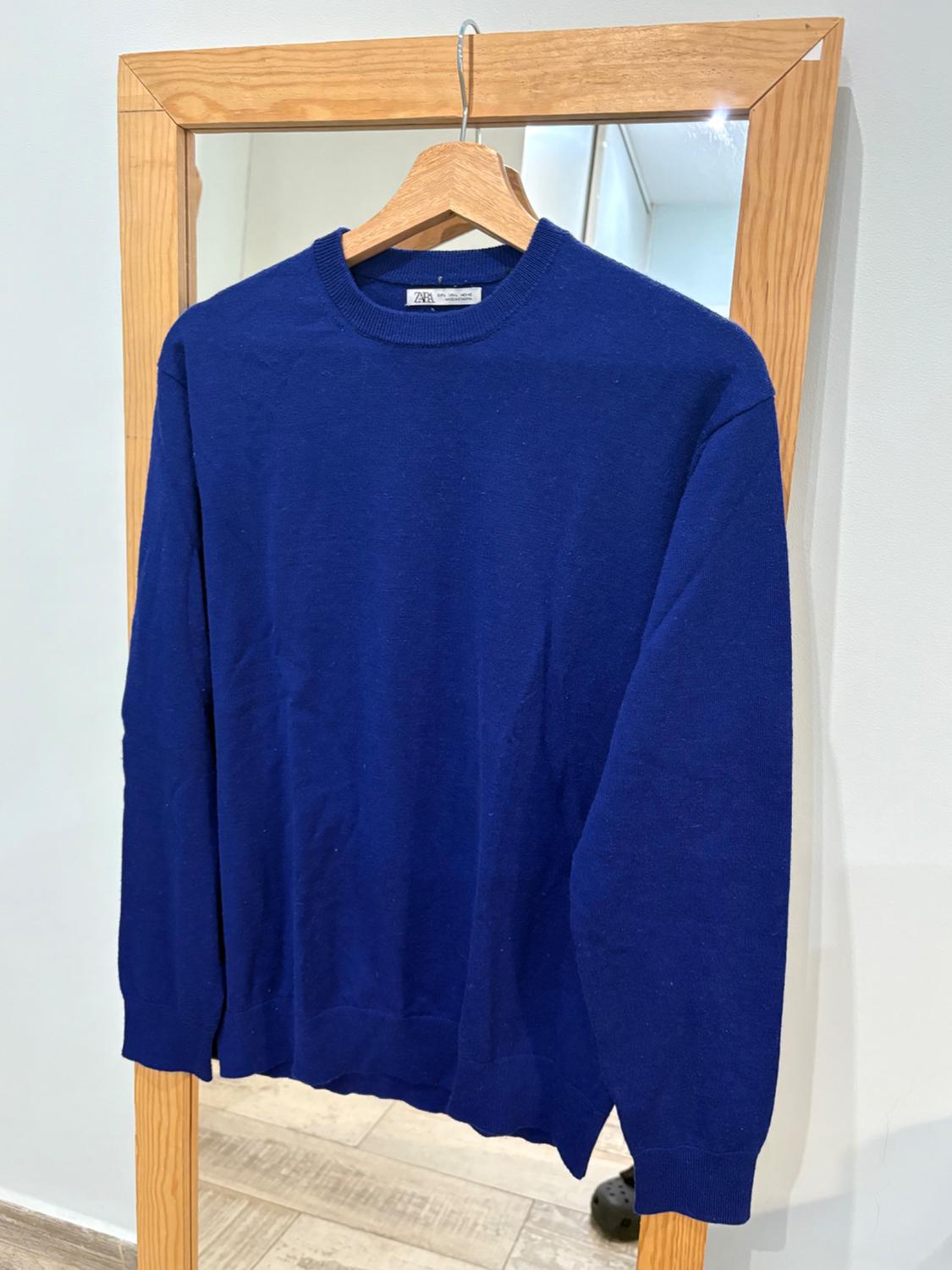 Blue jumper hanging on a wooden hanger in front of a mirror. Label visible on the neck. White background and light wooden floor.