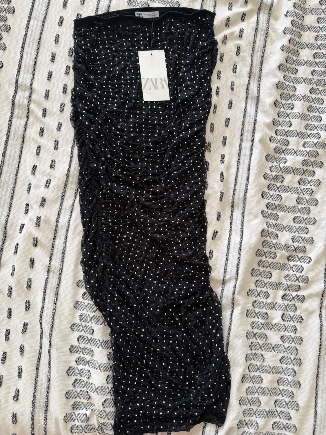 Black gathered skirt with white dots and ZARA label, laid out on a white blanket with a black geometric pattern. Relaxed style.