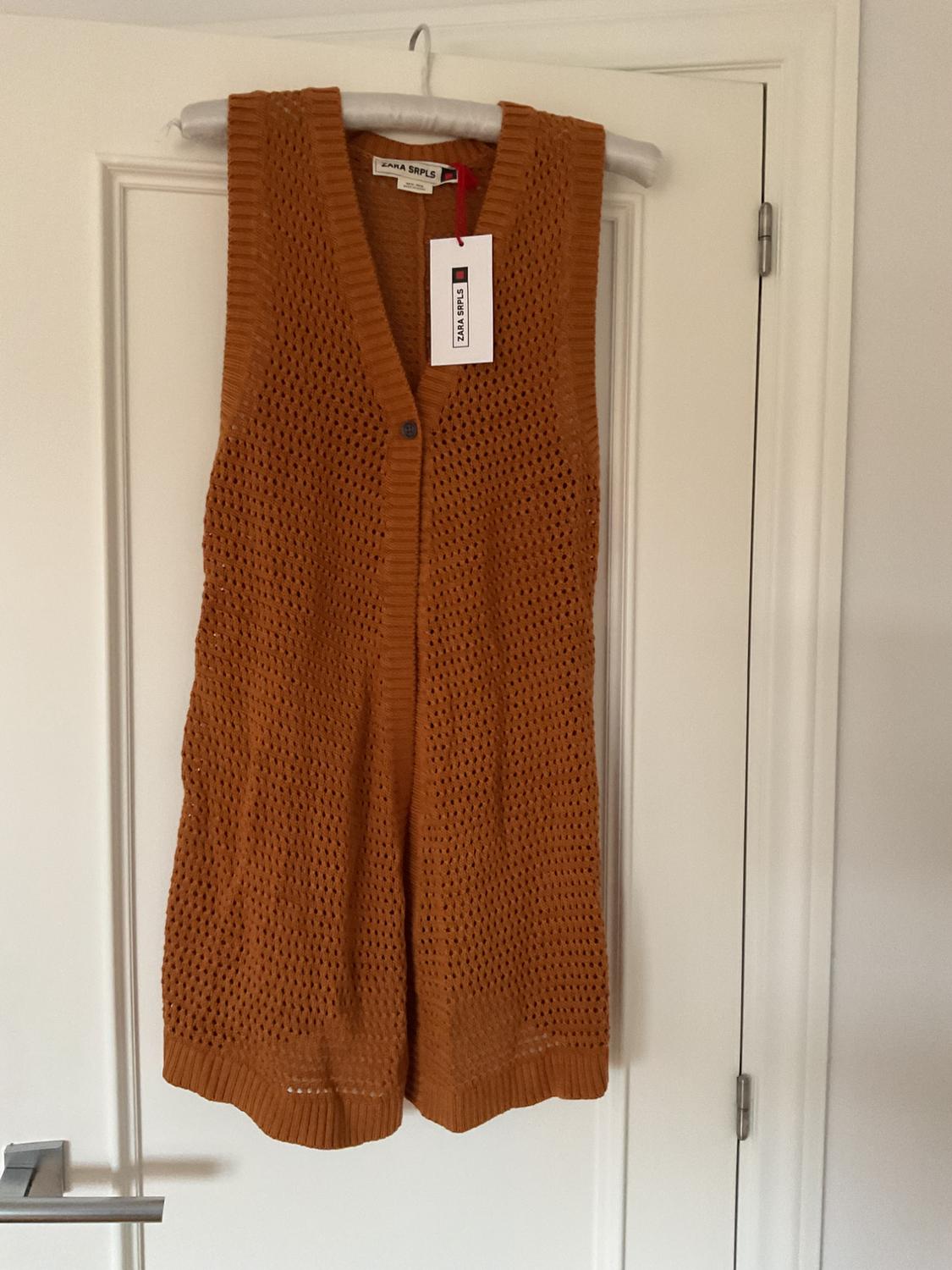 Brown knitted waistcoat hanging on a hanger over a white door. Visible label with the text "ZARA SRPLS".