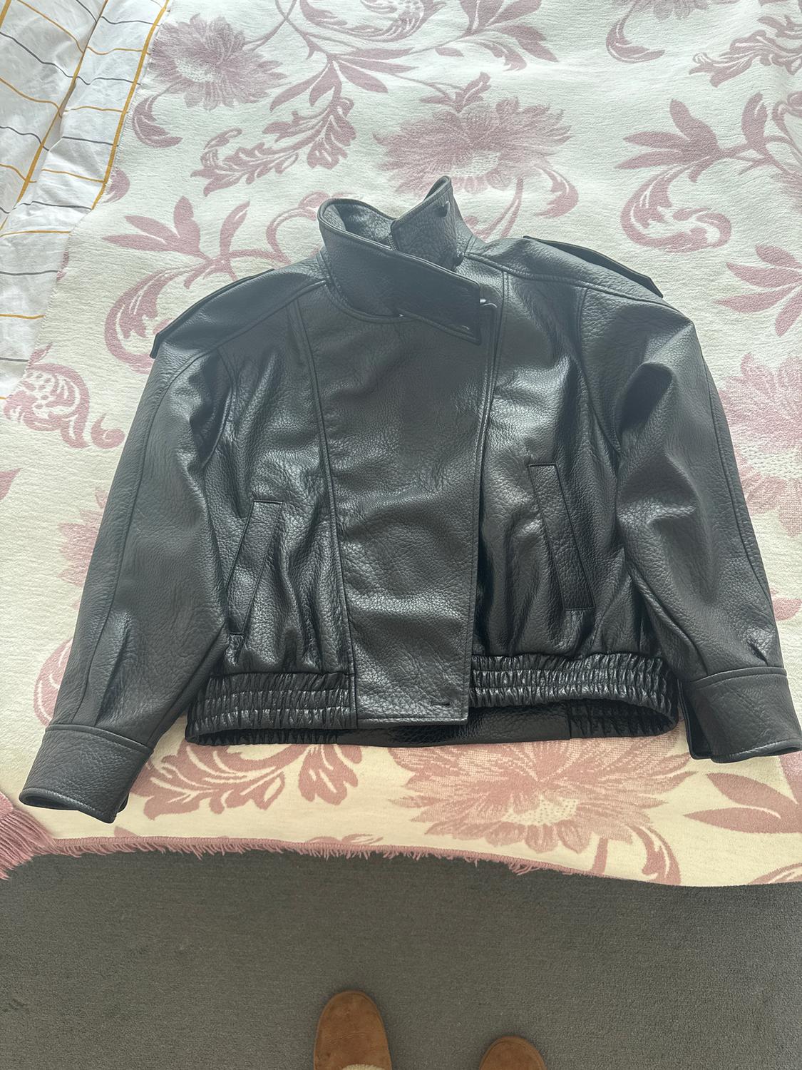 Black leather jacket on a bed with a pink floral bedspread. Elasticated hem and high collar. Grey carpet background.