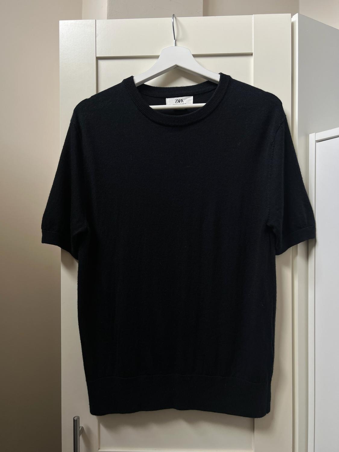 Black short-sleeved knitted jumper hanging on a white hanger in front of a white wardrobe. The label reads ZARA.