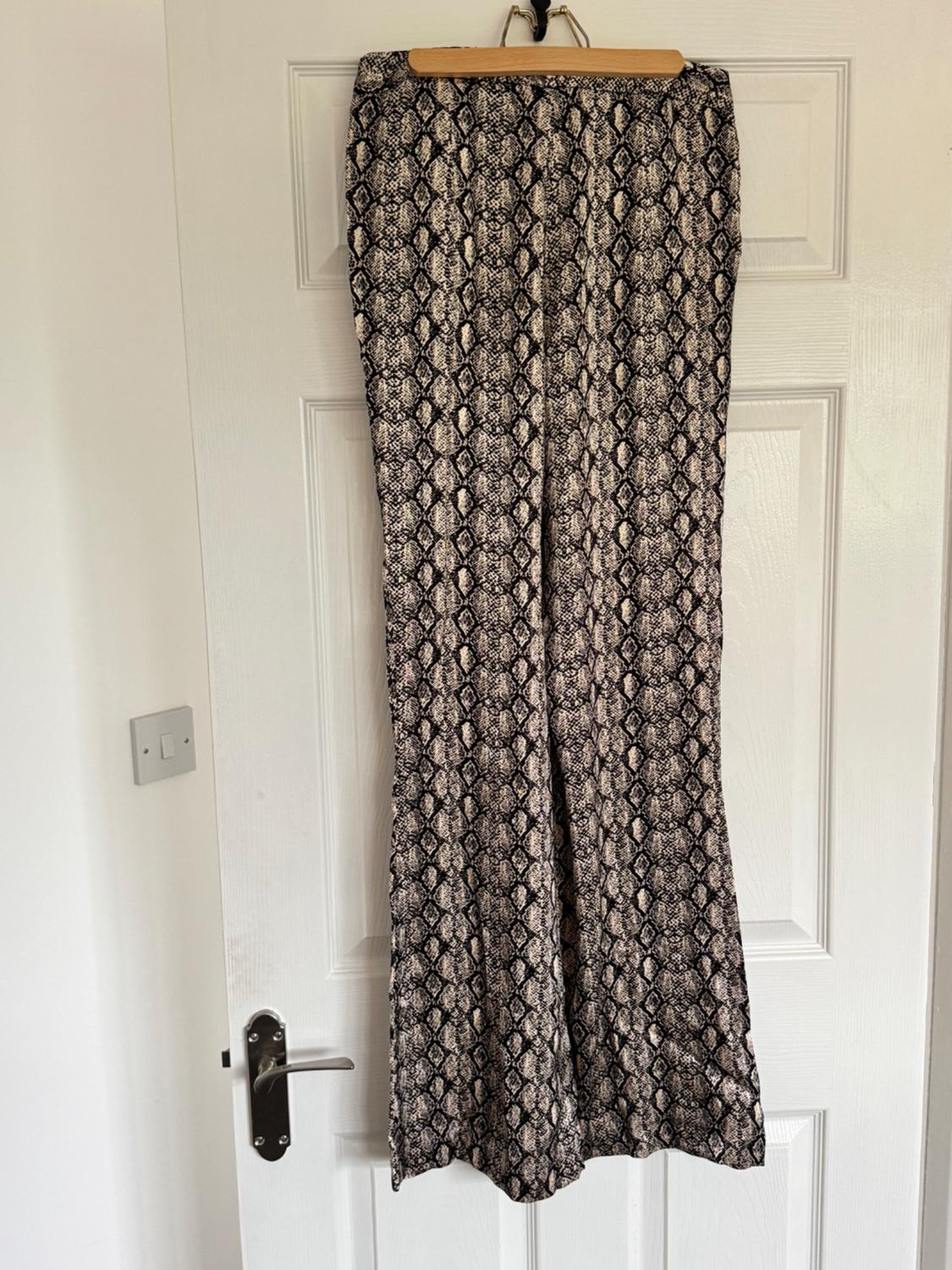 Snake print trousers in beige and black tones, hanging on a hanger over a white door with a silver knob.