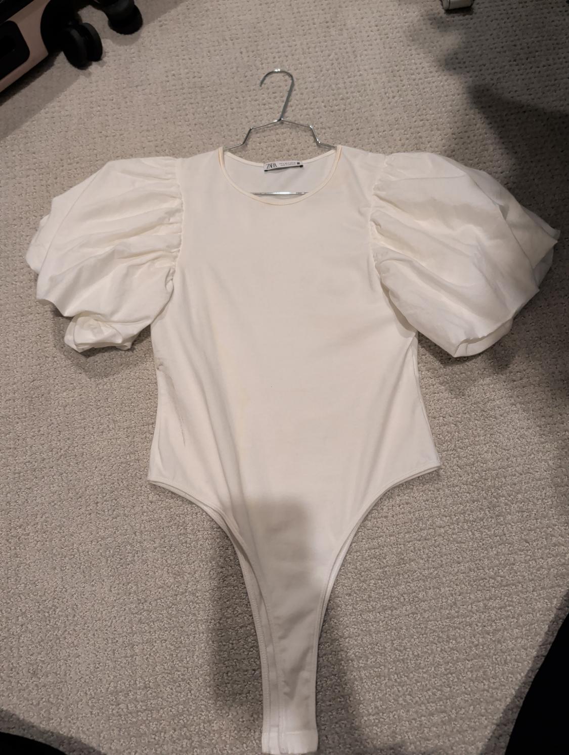 White bodysuit with puff sleeves on a hanger on a beige carpet. Label visible on the collar.