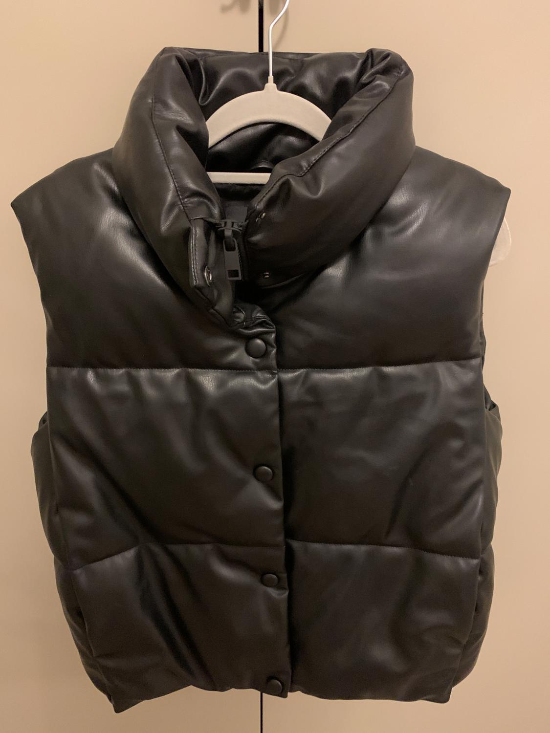 Black padded gilet hanging on a white hanger against a beige background. Design with buttons and a high collar.