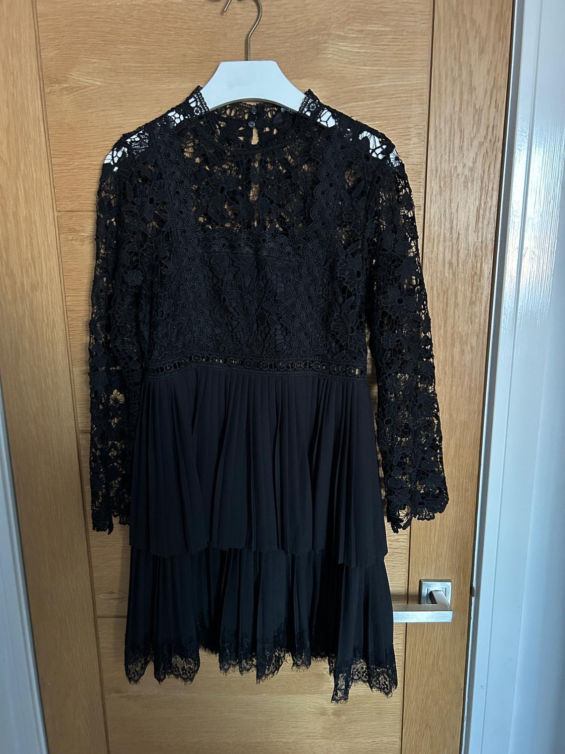 Black lace dress with long sleeves and a pleated skirt, hanging on a white hanger over a light wood door.