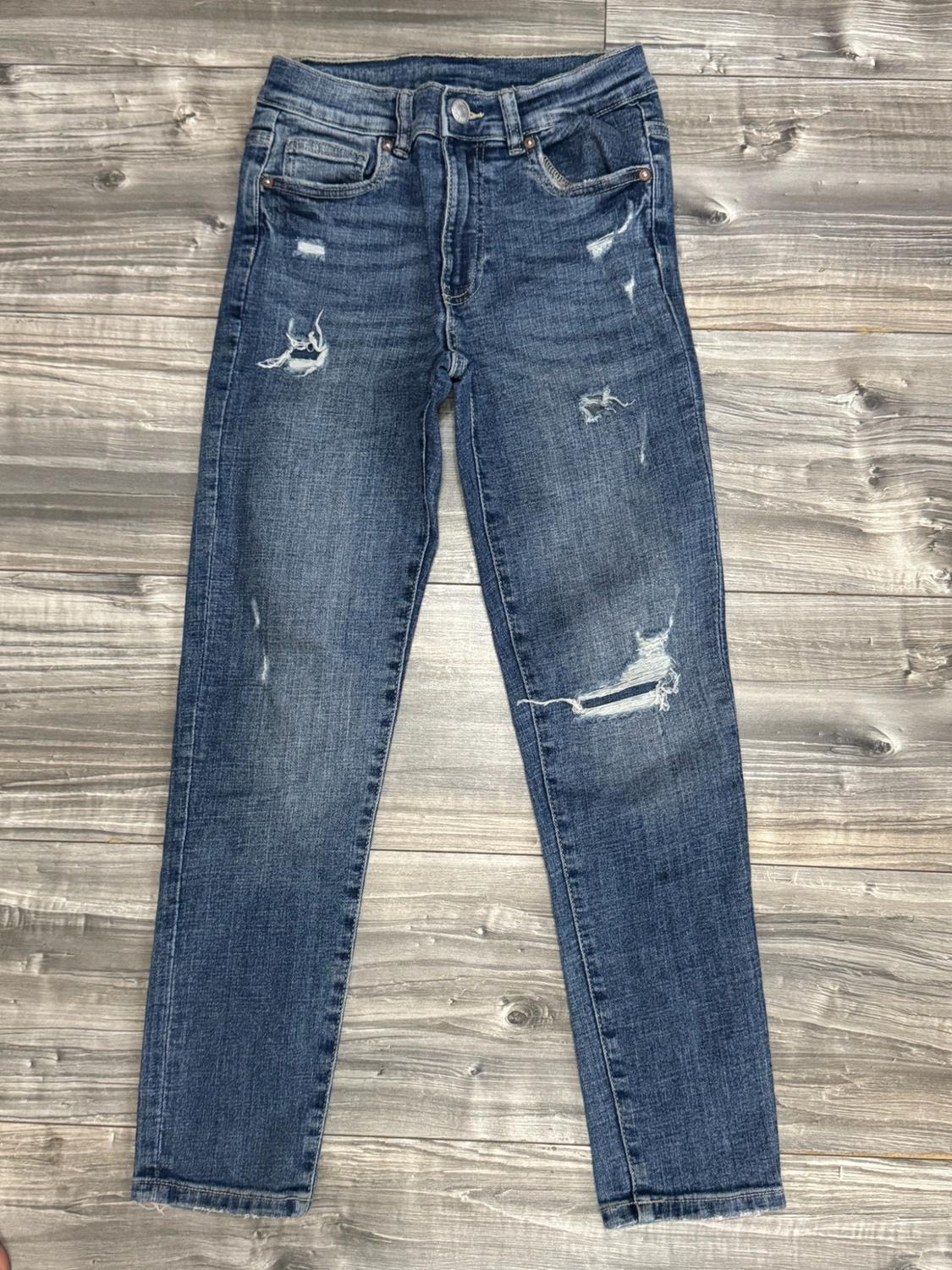 Blue denim trousers with distressed and ripped details, laid out on a wooden floor.