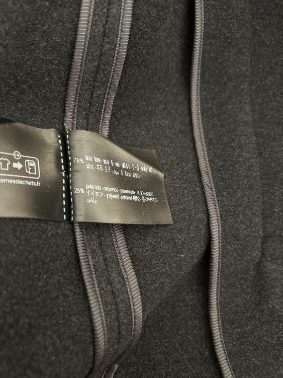 Black fabric label with multi-language text on material composition, sewn onto a dark-coloured garment with visible stitching.