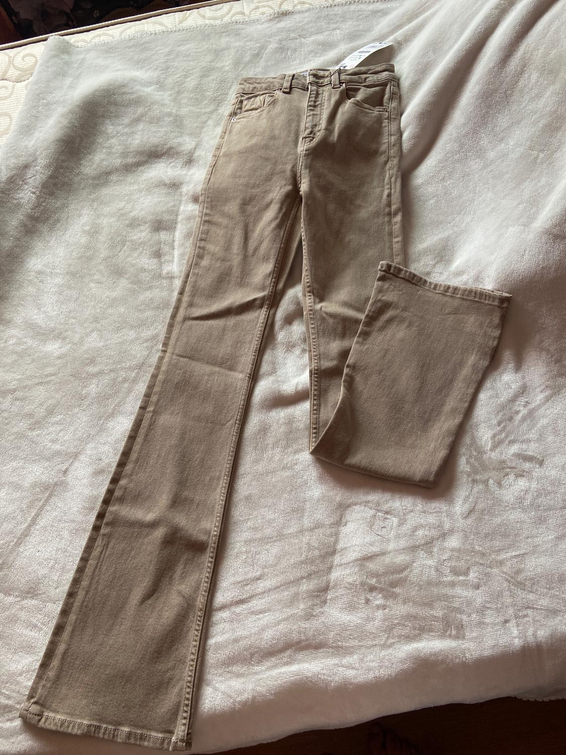 Beige flared trousers on a white blanket, with visible label on the waistband. Relaxed and casual atmosphere.