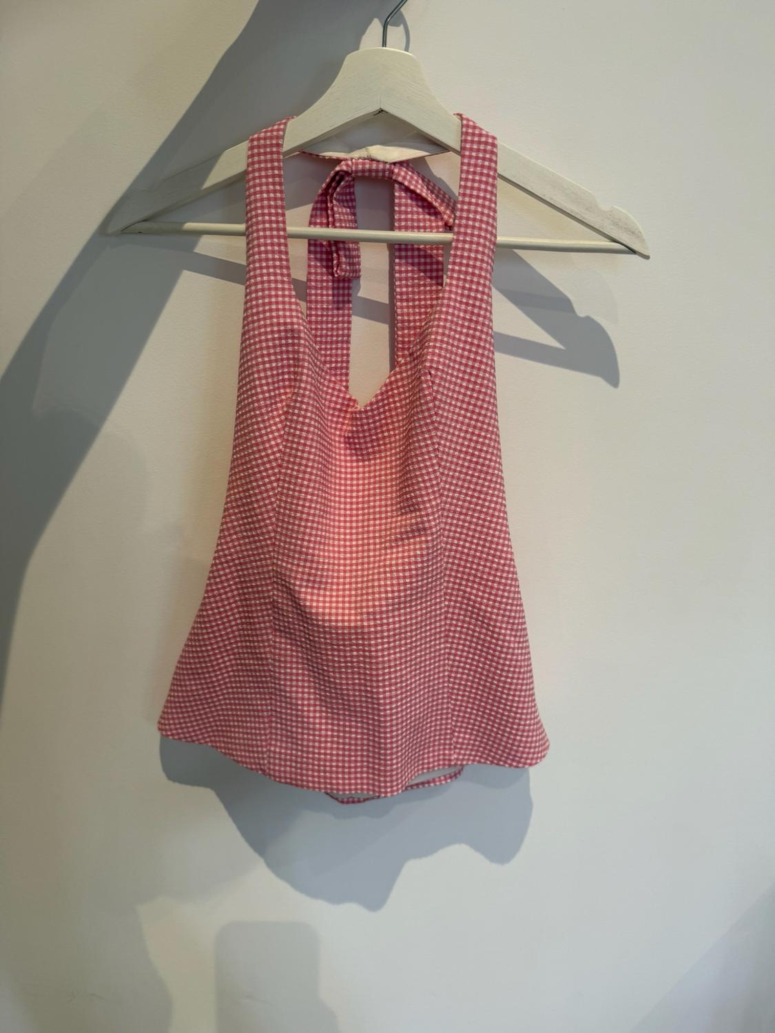 Top with pink and white checks and a halter neck hanging on a white hanger against a white wall with cast shadows.