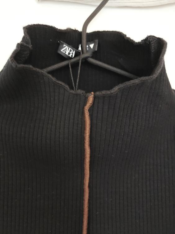 Black ribbed top hanging on a hanger, with visible central brown stitching. "ZARA" brand label on the inside of the neck.