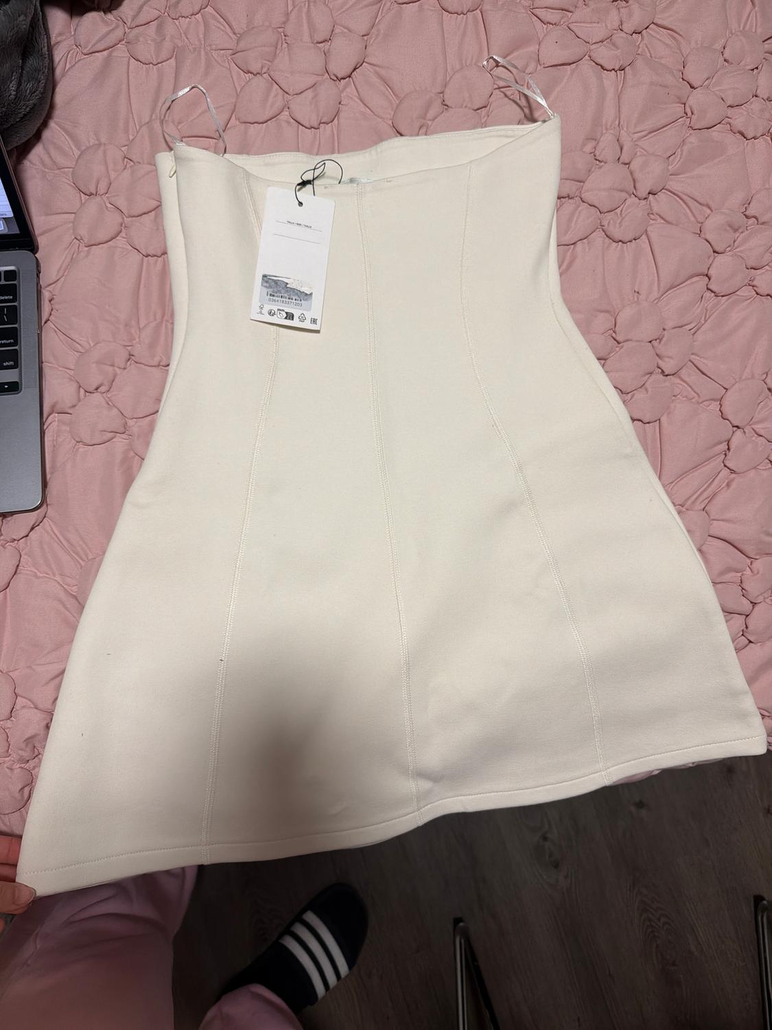 White strapless dress on pink bedspread, with hangtag. Part of a laptop and foot with sock visible.