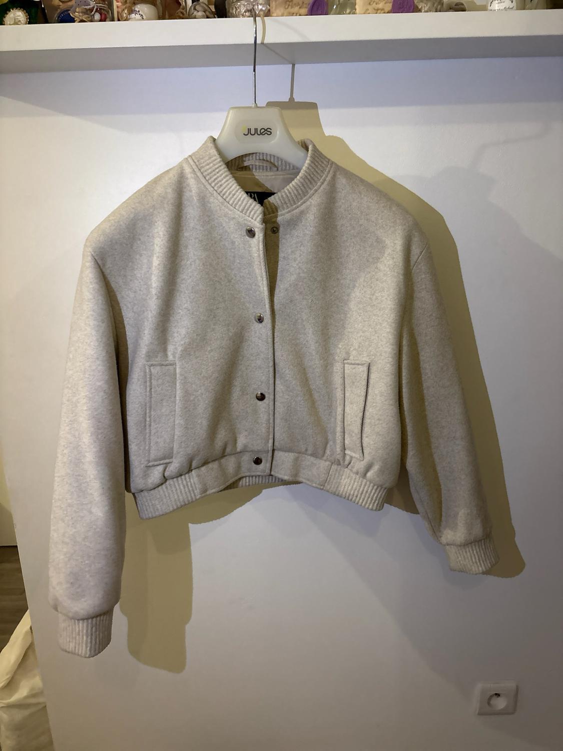 Light beige cropped bomber jacket with press studs and pockets, hanging on a hanger with the text JULES in front of a white wall.