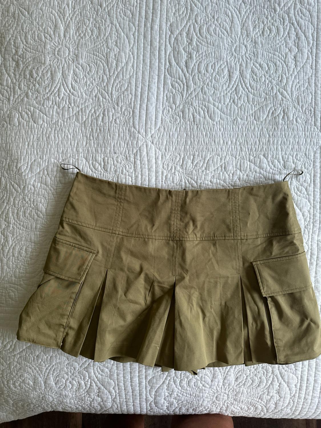 Pleated olive-green miniskirt with side cargo pockets, spread out on a textured white bedspread.