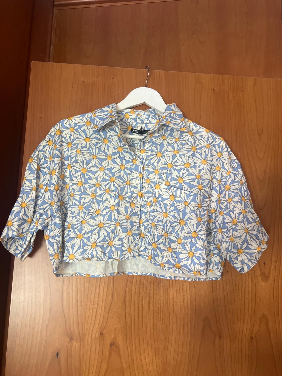 Short-sleeved crop top with white and yellow daisy print on a light blue background, hanging on a white hanger.