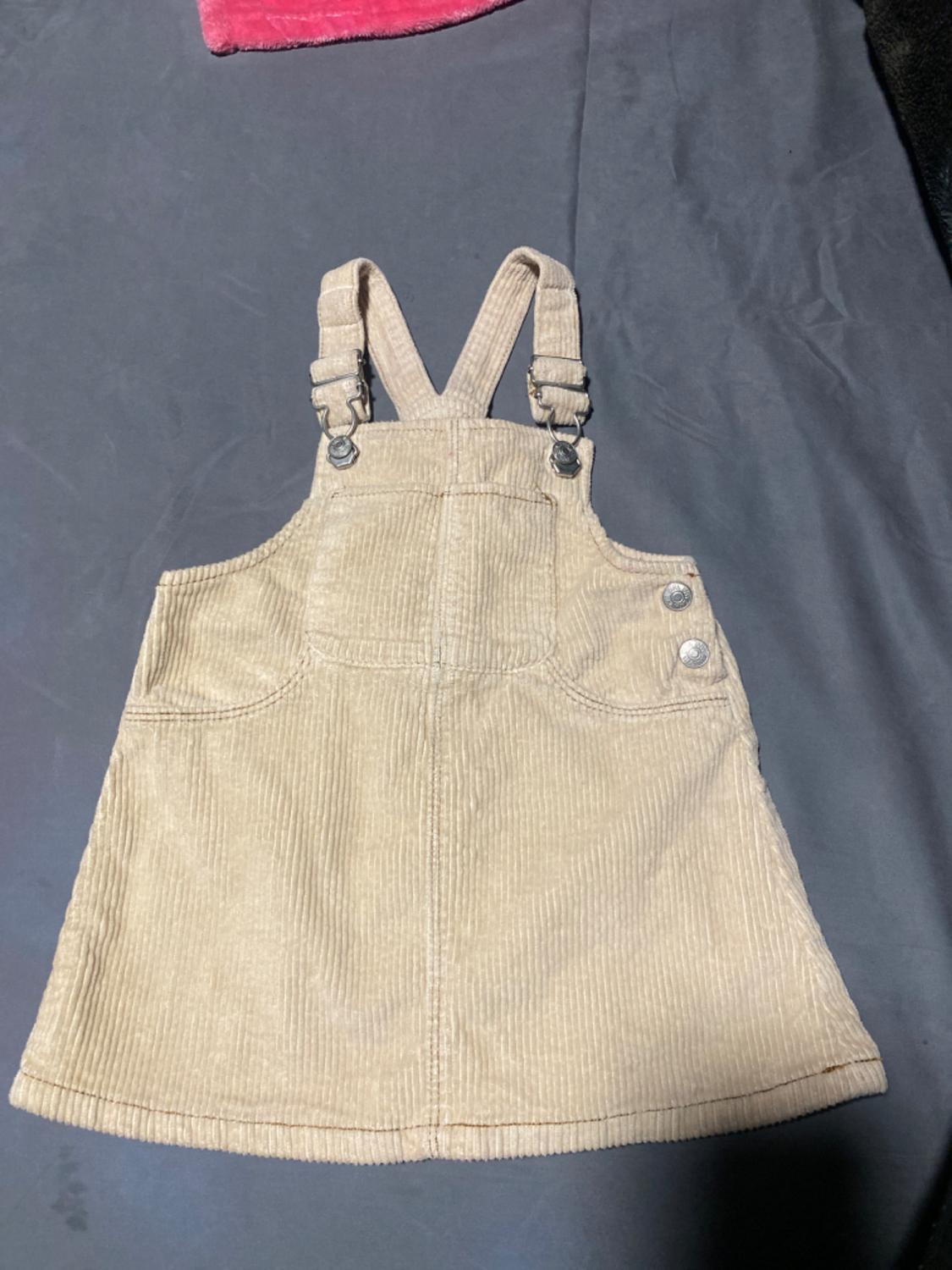 Beige corduroy dress with adjustable straps and buttons on the chest, on a grey surface. Part of a red fabric visible above.