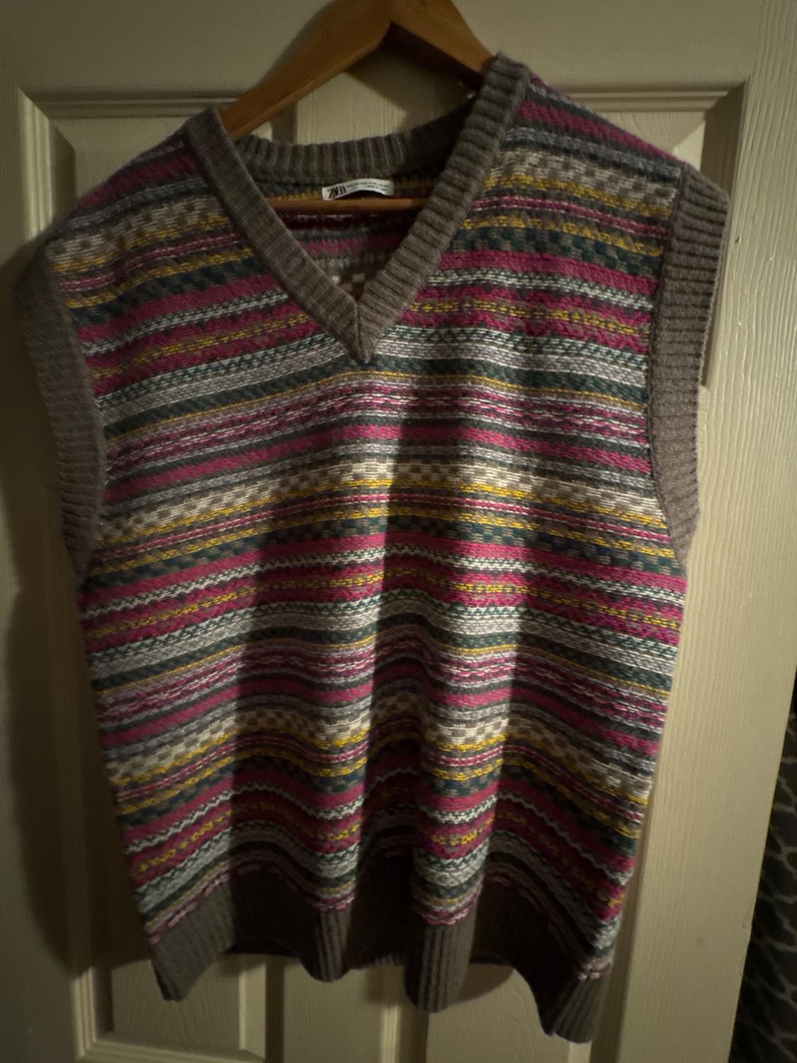 Multicoloured knitted waistcoat with horizontal stripes in pink, yellow, and grey tones, hanging on a wooden hanger.