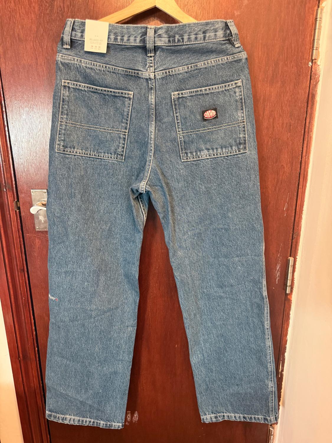Blue jeans hanging on a hanger over a wooden door. Rear view with pockets, patch, and label with 'RELAXED FIT' text.