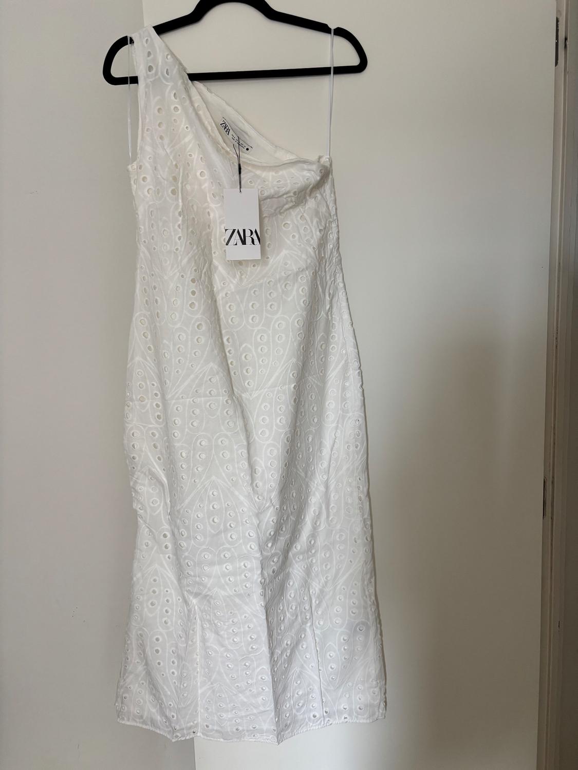 One-shoulder white dress with eyelet embroidery, hanging on a black hanger. Visible label with the text "ZARA".