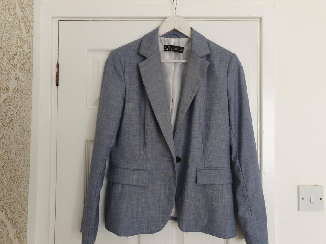 Grey-blue blazer with a ZARA tag hanging on a hanger on a white door. Relaxed home atmosphere with wallpaper.