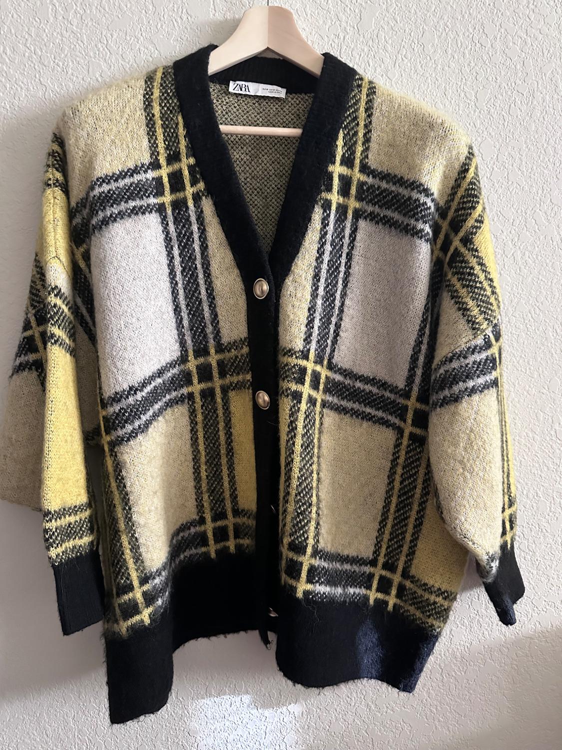 Large check cardigan in shades of yellow, black and white, hanging on a wooden hanger against a white wall.