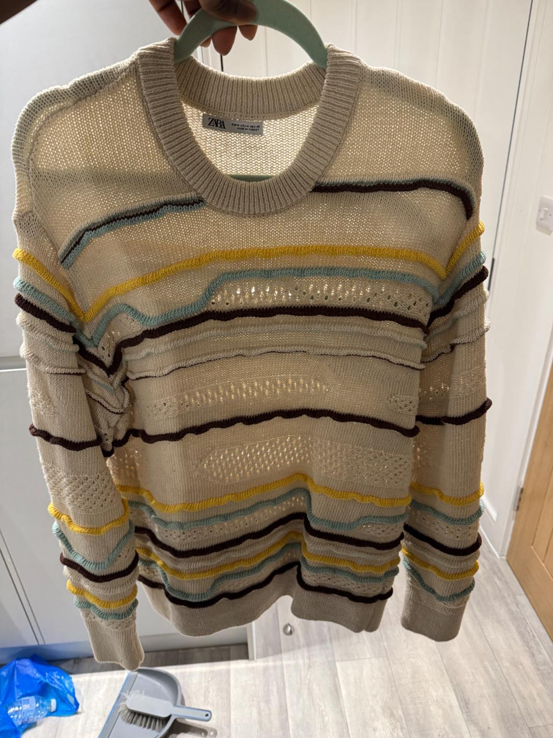 Beige knitted jumper with horizontal stripes in brown, yellow, and light blue, hanging on a green hanger indoors.
