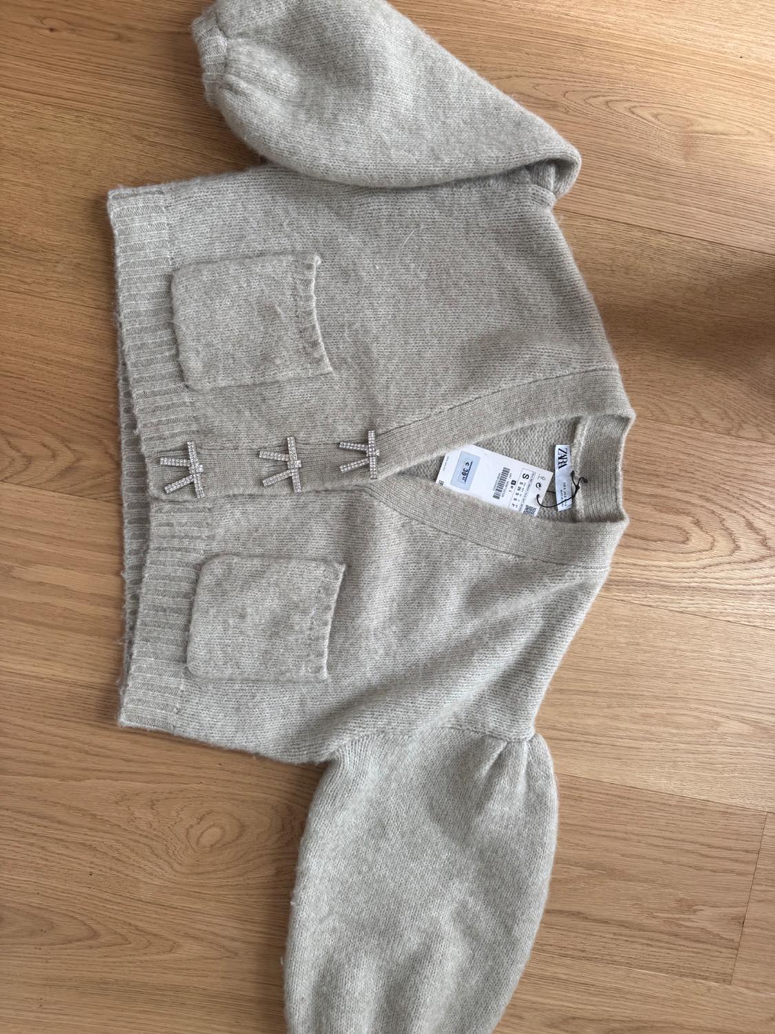 Light grey knitted jacket with puffed sleeves, front pockets, and sparkly snap fasteners, on a wooden floor.