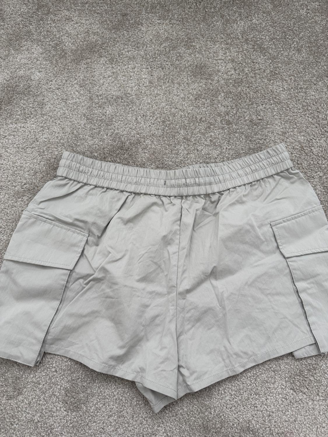 Grey shorts with large side pockets, placed on a grey rug. Wrinkled fabric and elasticated waistband.