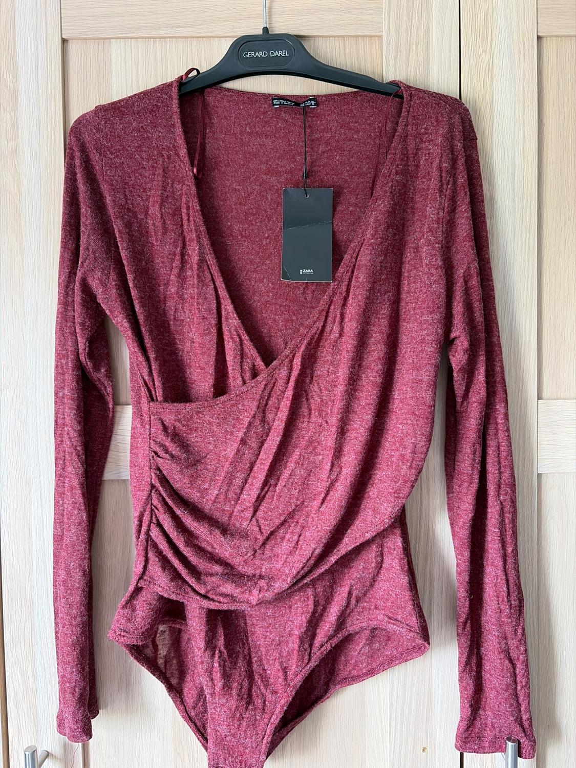 Long-sleeved maroon bodysuit with a wrap neckline and ZARA label, hanging on a wooden door on a black hanger with the text GERARD DAREL.