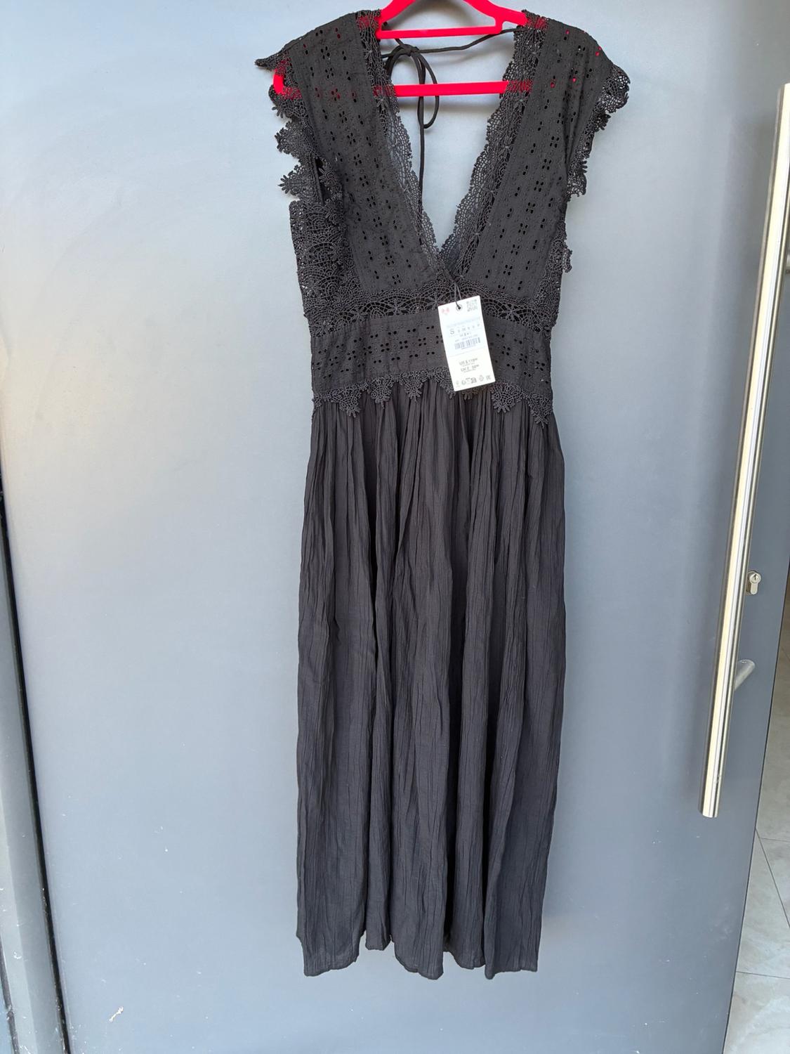 Long black dress with a V-neckline, lace details and a textured skirt, hanging on a fuchsia hanger over a grey door.