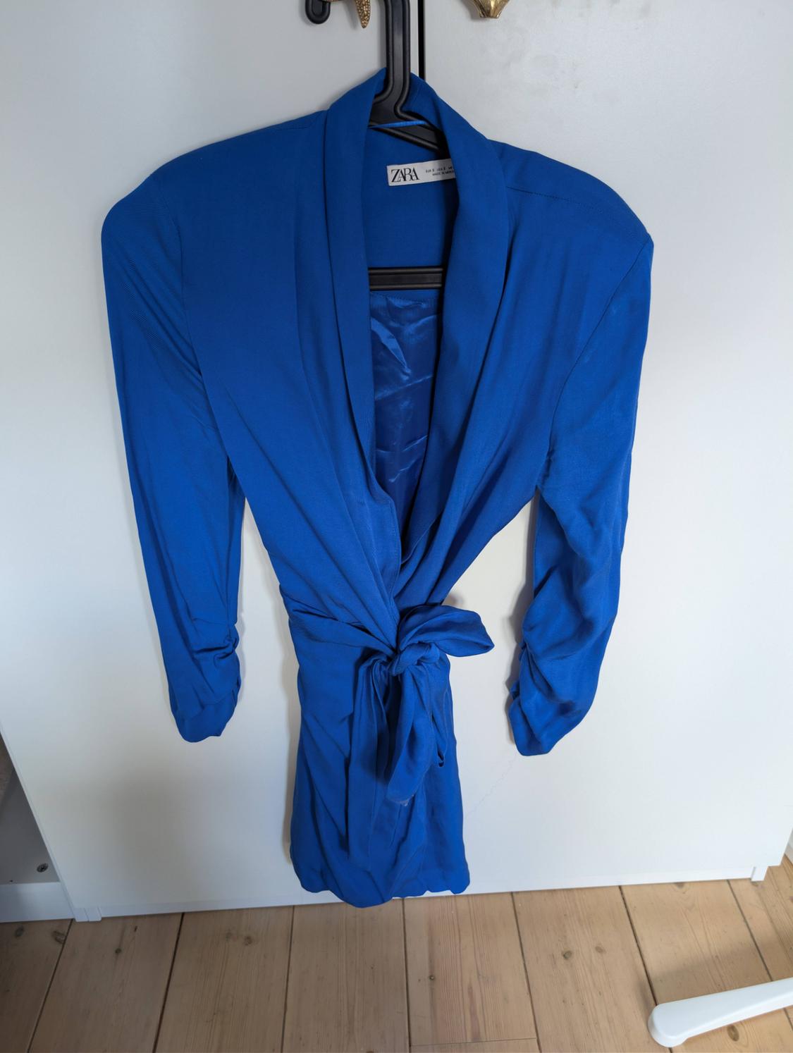 Blue blazer with a knotted belt hanging on a black hanger in front of a white wardrobe and a wooden floor. Text: ZARA.