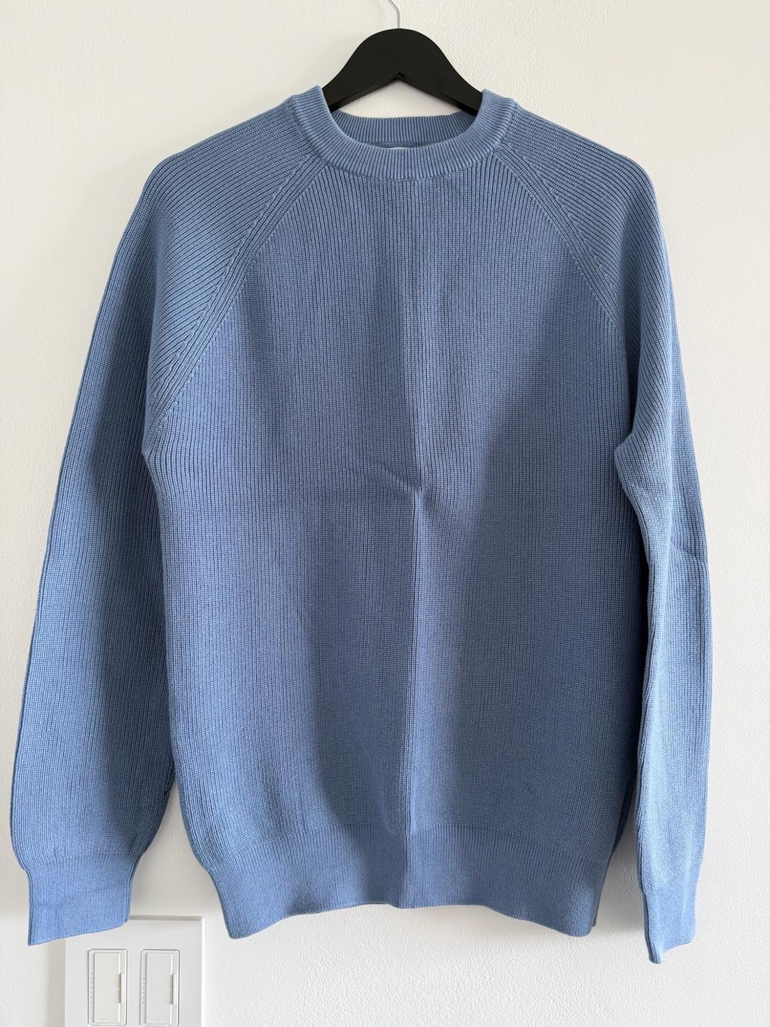 Light blue knit sweater with a crew neck, hanging on a black hanger against a white wall.