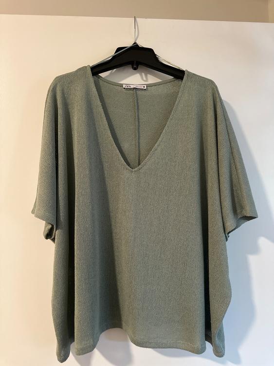 Olive green knit blouse with a V-neck, hanging on a black hanger against a white background. Visible tag on the collar.