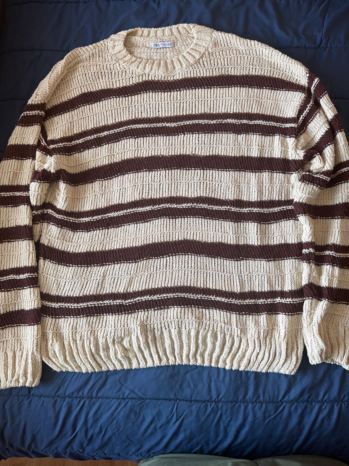 Beige knitted jumper with horizontal brown stripes on a blue surface. Visible label on the collar.
