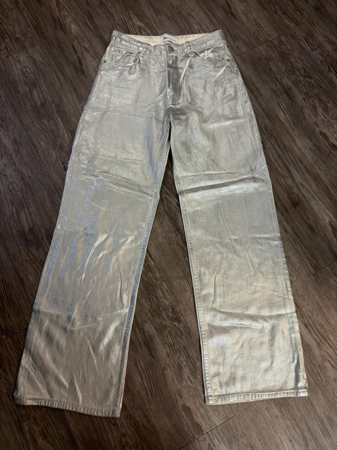 Long, wide-leg trousers with a silver metallic finish, laid out on a wooden floor.
