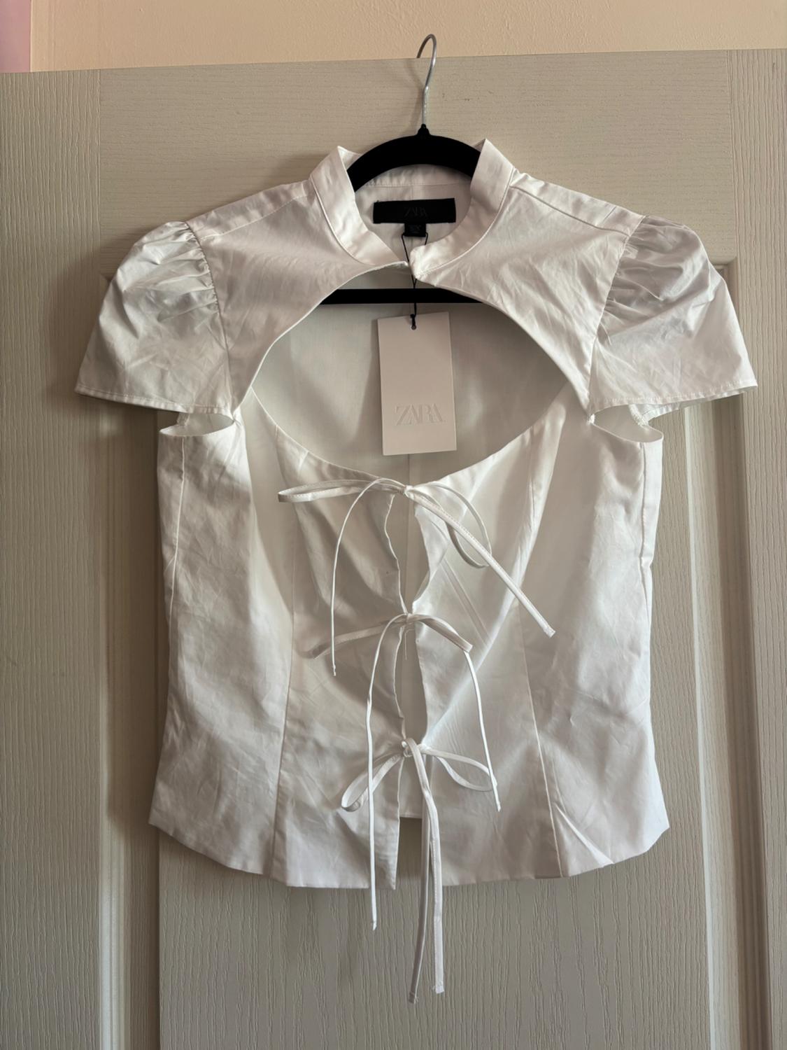 White top hanging on a door. Short-sleeved design, high neck, chest opening and tie fastening. Label with ZARA text.
