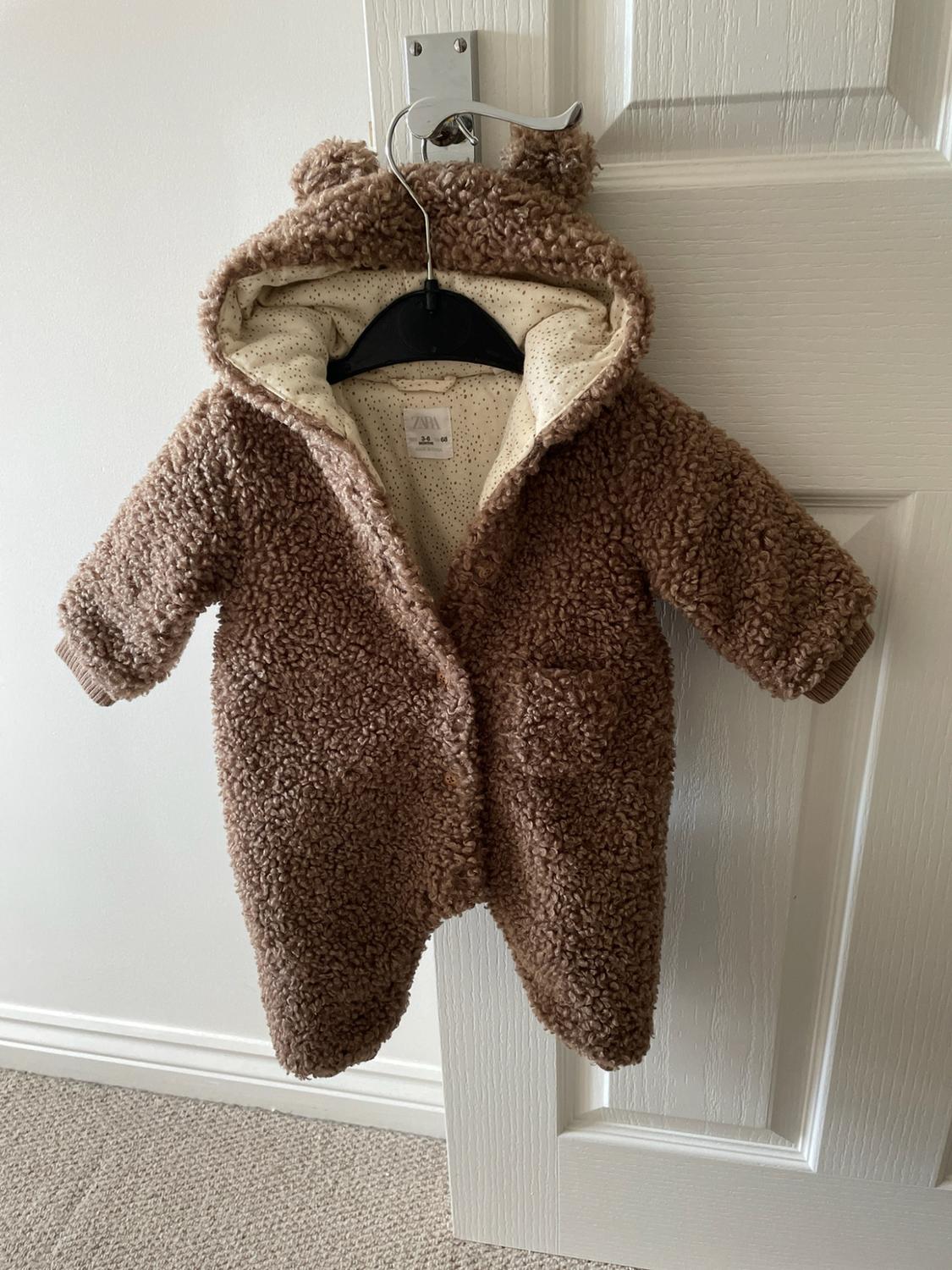 Brown teddy-textured baby all-in-one with hood and ears, hanging on a white door. ZARA label visible inside.