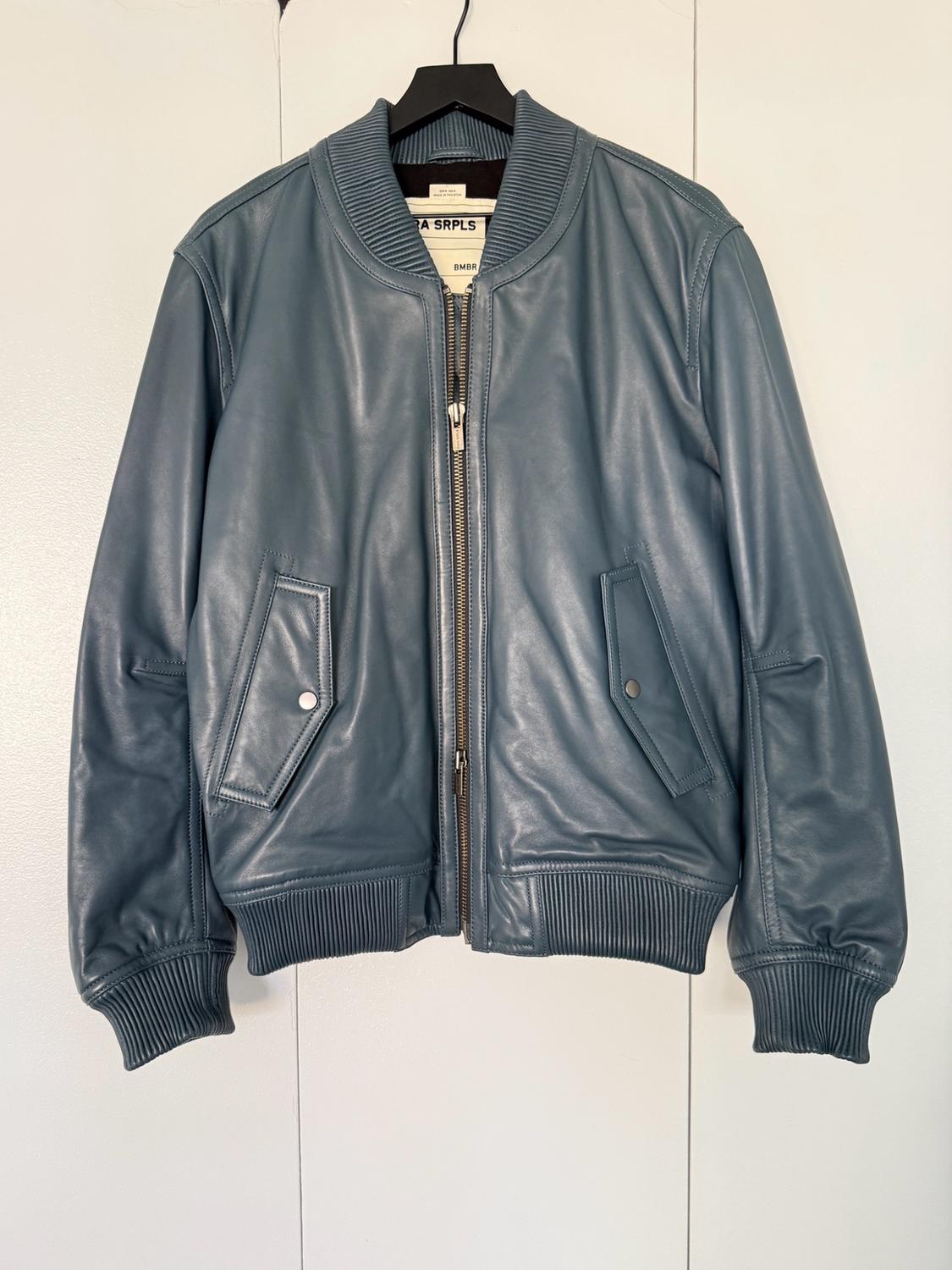 Blue bomber jacket with zip and pockets, hanging on a hanger against a white background. Interior label with the text ZARA SRPLS BMBR.