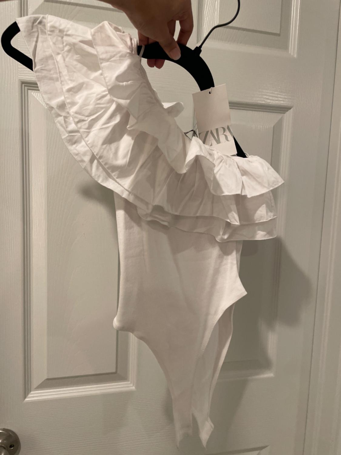 White bodysuit with frills on the shoulders, hanging on a black hanger in front of a white door. Label visible with the text "ZARA".