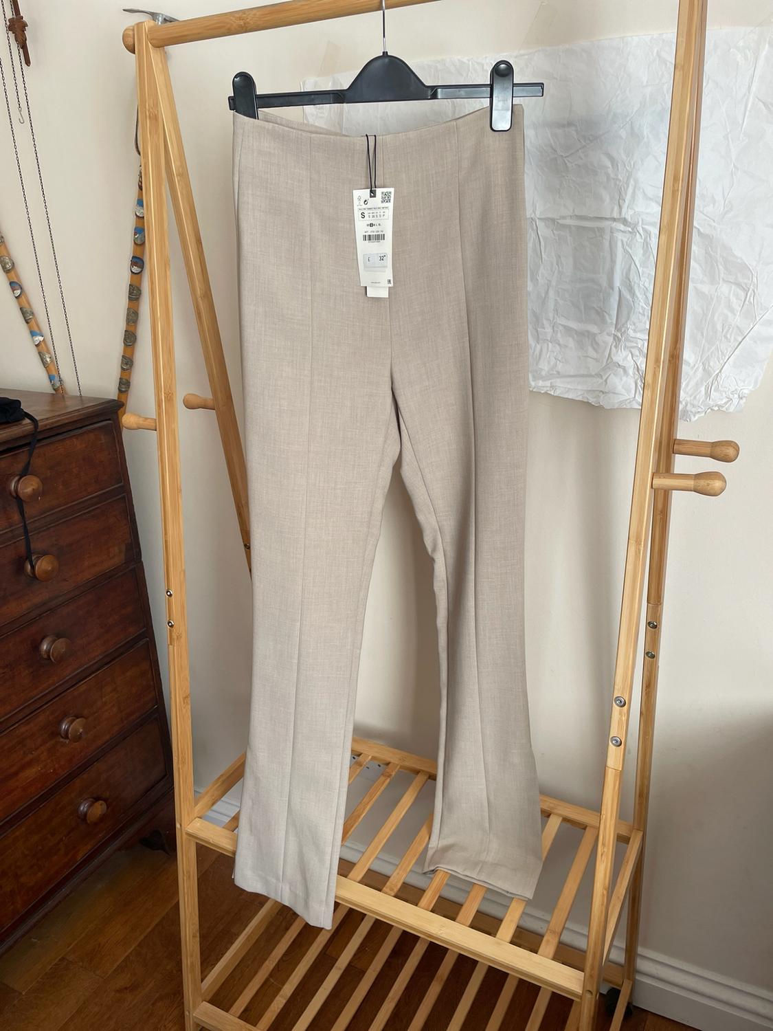 Beige trousers with a front seam and label, hanging on a wooden clothes rail. In the background, a classic chest of drawers and a light-coloured wall.